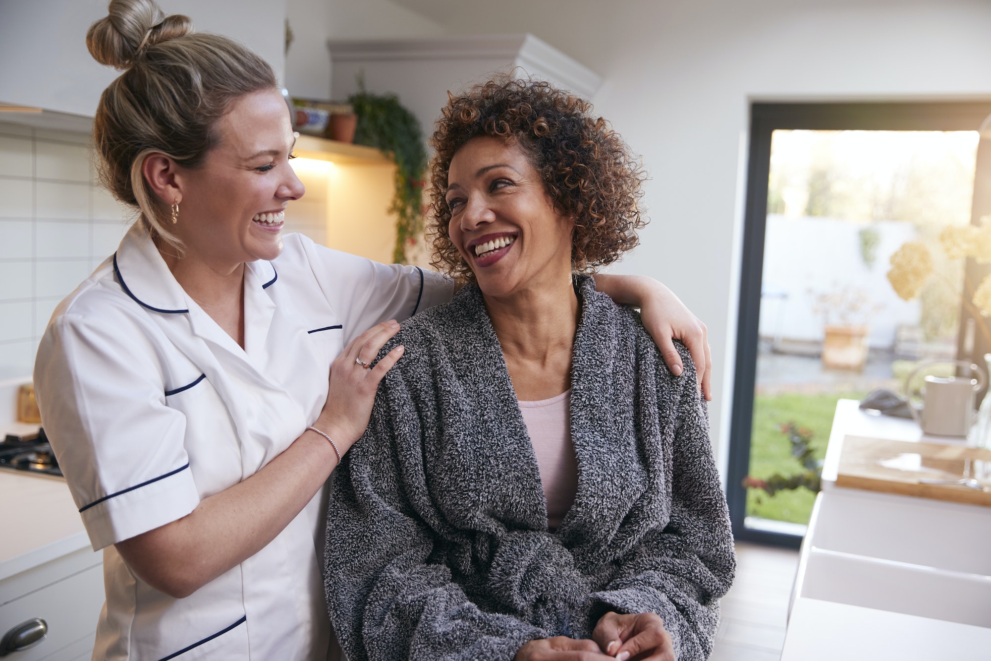 Mature Woman In Dressing Gown Talking With Female Nurse In Kitchen At Home